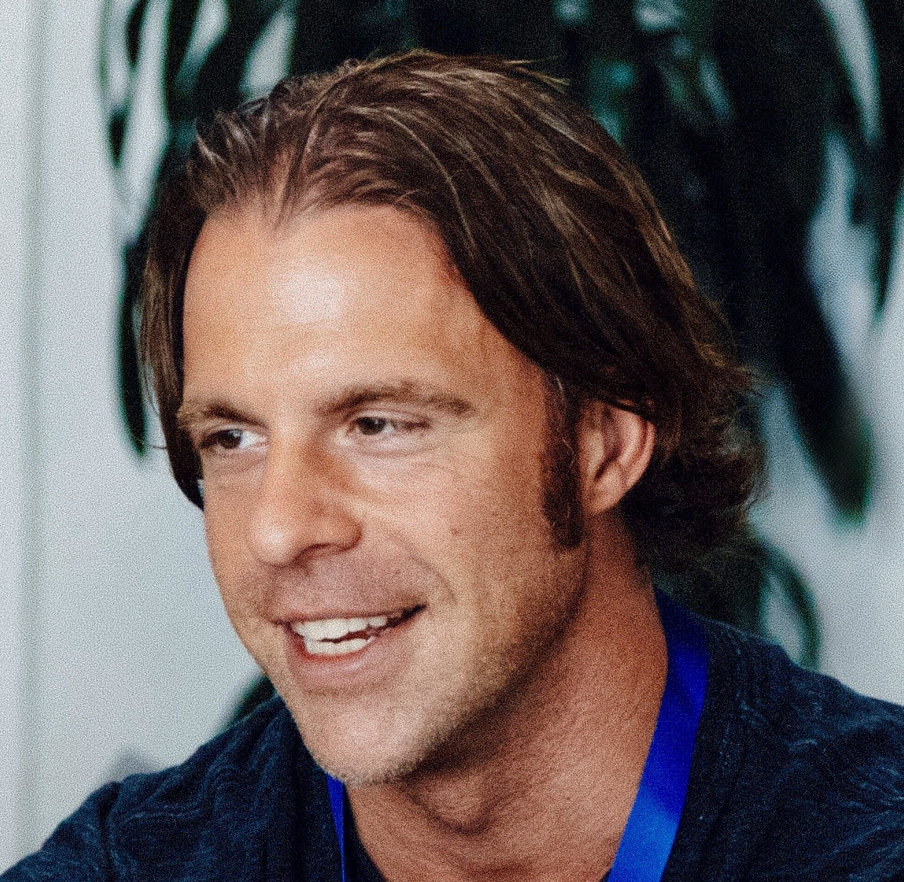 Smiling man with medium-length brown hair and a blue lanyard, seated indoors with blurred greenery in the background.